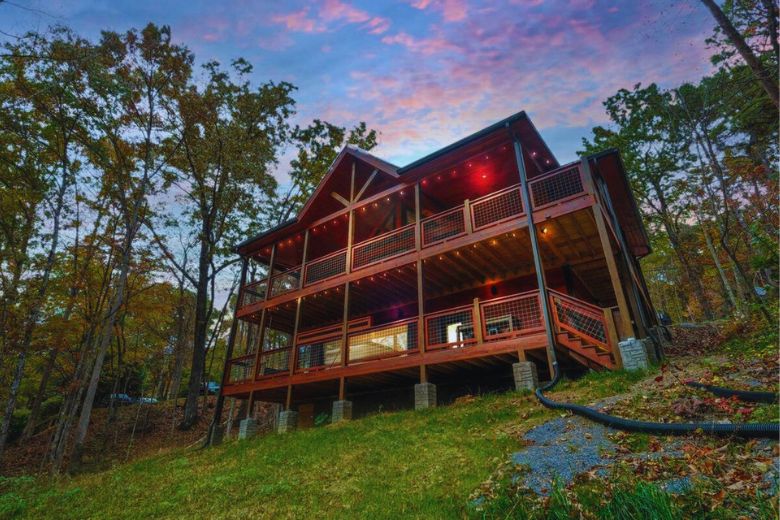 Large two-story cabin with wraparound decks and string lights glowing at dusk, nestled on a wooded hillside with a grassy lawn and surrounded by tall trees under a colorful sunset sky.